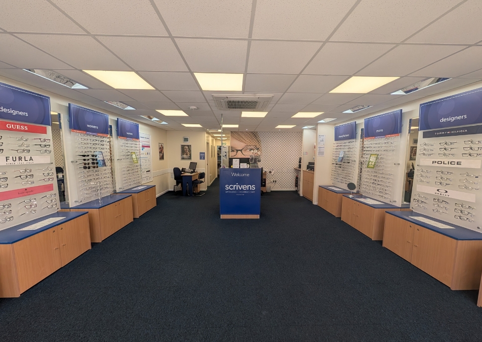 Interior of an optical store with wall-mounted display racks of glasses on both sides and a blue reception desk in the center.