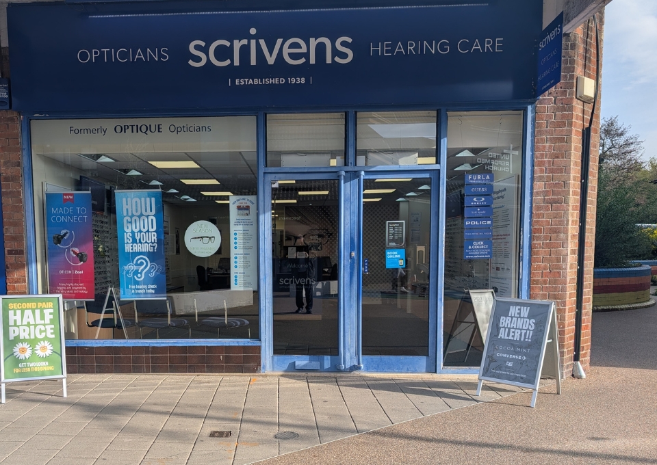 Blue storefront of Scrivens Opticians Hearing Care with glass doors, window posters, and sidewalk signs; advertisements for half-price deals and new brands visible outside on a sunny day.