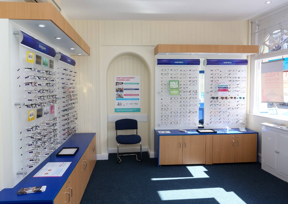 Eyewear shop interior with wall displays full of eyeglasses and a blue cushioned chair by the wall.