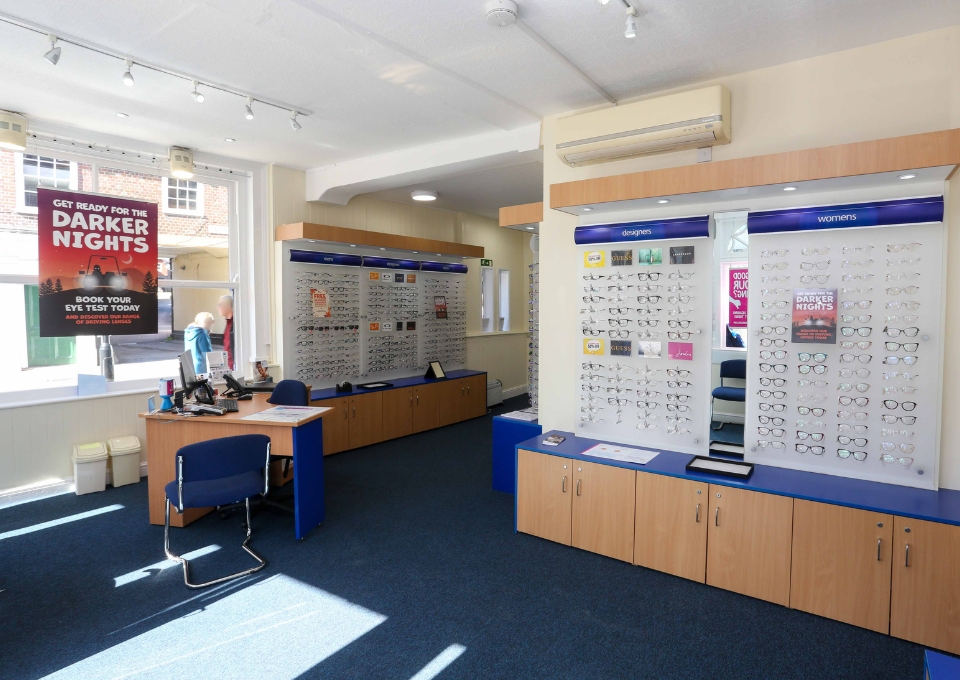 Interior of an eyeglasses store with wall displays of frames, a reception desk, and a poster about darker nights near a window, bright and welcoming.