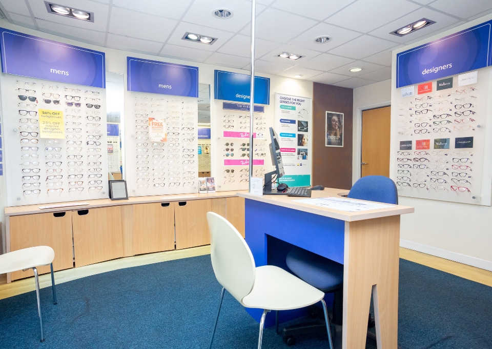 Optician showroom interior with wall displays of glasses labeled 'mens' and 'designers', a reception desk with computer, and seating area.