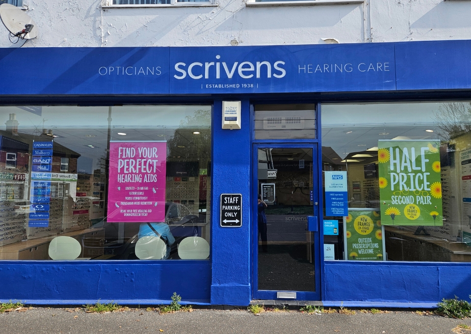 Blue storefront for Scrivens Opticians Hearing Care, with large glass windows displaying pink and green promotional posters.