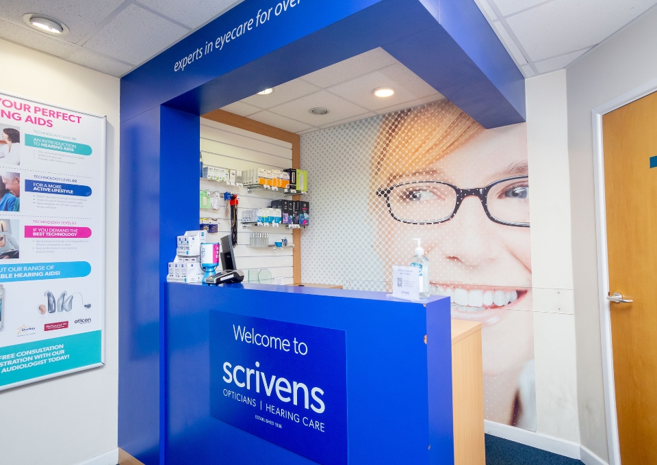 Reception desk at Scrivens Opticians with blue signage and a large wall graphic of a smiling person wearing glasses; welcome sign visible on the counter.