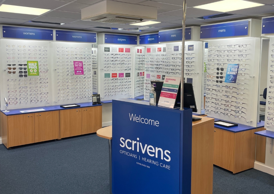 Shop interior of an eyewear store with rows of eyeglasses on display behind a blue Welcome Scrivens desk. Signage for womens, mens, designers visible on walls.