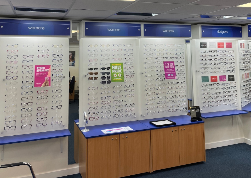 Wall display of women's eyeglasses in a store, with multiple racks and blue 'womens' signs above.