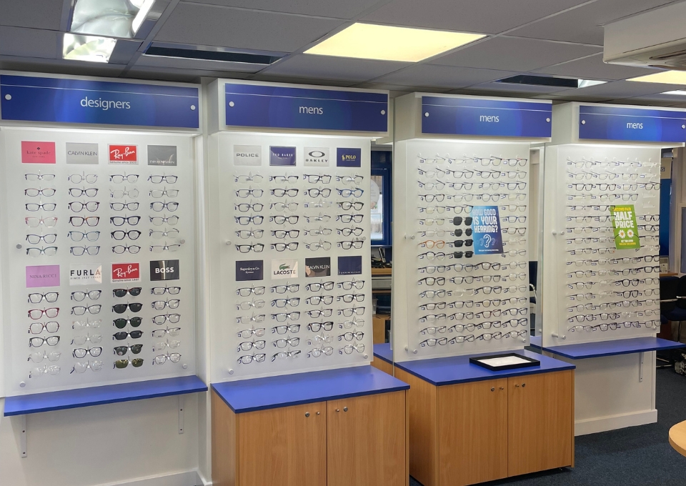 Rows of eyeglasses on display panels labeled designers and mens, arranged on white boards with blue accents in a store showroom.