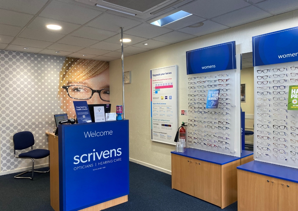 Reception desk with Scrivens branding in an eyewear shop; wall of women's glasses displayed on the right.