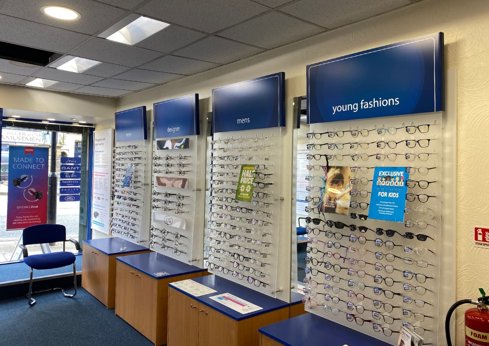 Interior of an eyewear store with wall-mounted glasses displays labeled men's and young fashions, blue signage, and wooden cabinets below the displays.