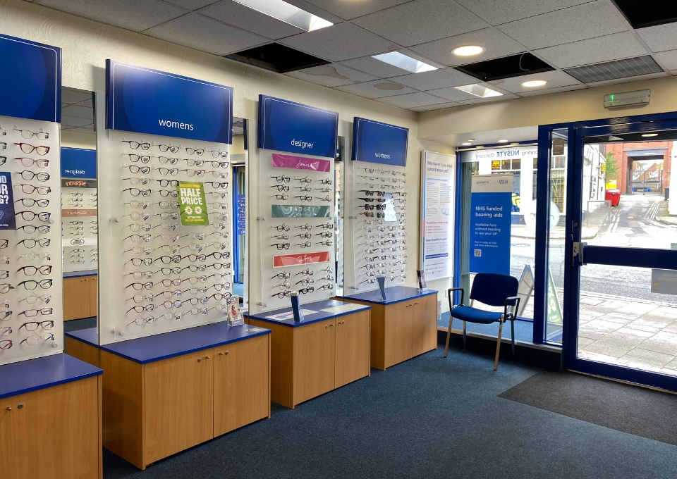 Eyewear display with rows of glasses labeled women's and designer inside a shop near the entrance.','Interior of eyewear store showing wall-mounted frames, blue signs, and wooden cabinets.','Row of display stands with sunglasses and prescription glasses in a bright shop by the entrance.','Store entrance area with blue signage, chair, and glass doors looking out to the street.','Gently lit optical shop featuring multiple glass-frame displays and a seating area by the door.