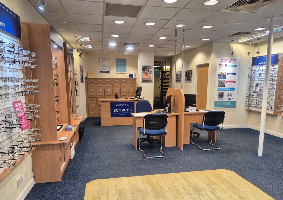 Interior of an optician store with wall eyewear displays on the left, a reception desk center, and blue chairs nearby.