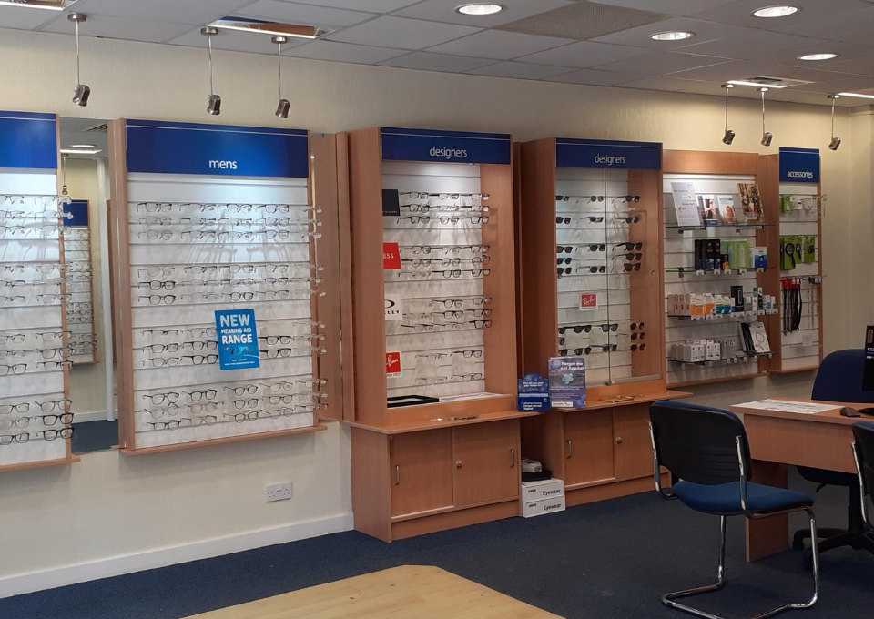 Eyewear store interior with wall-mounted glasses displays labeled 'mens' and 'designers' and a service desk nearby.