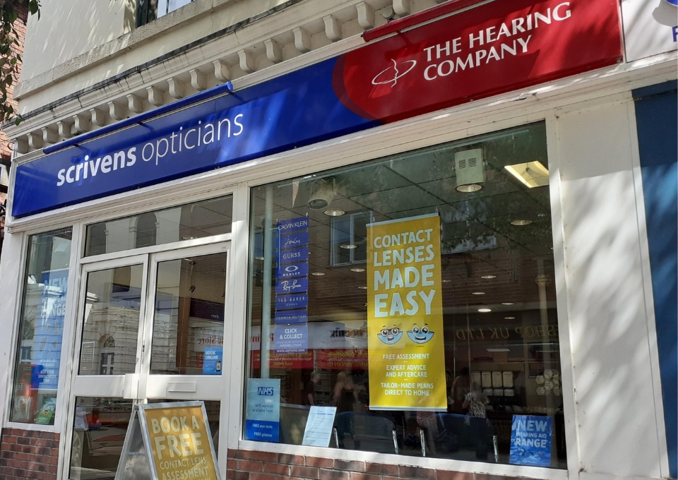 Exterior storefront for an optical shop with blue 'scrivens opticians' sign and red 'The Hearing Company' banner above the window. A yellow poster in the window reads 'Contact Lenses Made Easy.'