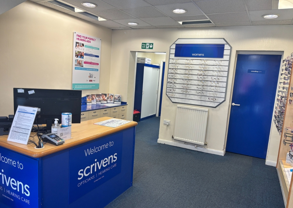 Interior of an optician's shop with a blue reception desk reading 'Welcome to Scrivens', eyewear on wall display, and a blue door nearby.