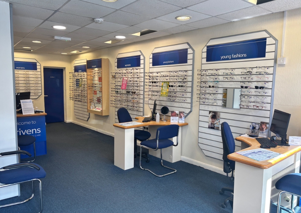 Eye care shop interior with wall displays of prescription glasses, labeled sections for women and young fashions, and service desks with computers and blue chairs.