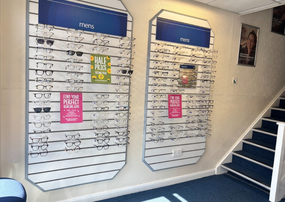Two wall-mounted displays of men's eyeglasses on horizontal rods, with blue 'mens' headers and promotional posters (e.g., 'Half Price' and 'Find your perfect hearing aids').