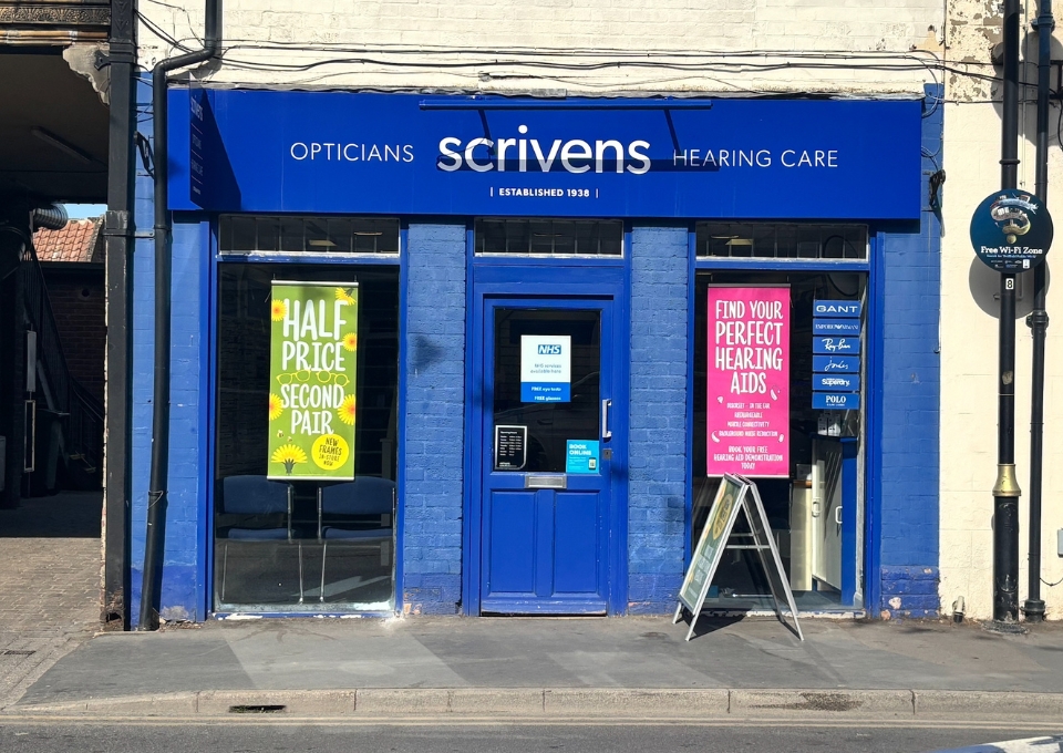 Blue storefront of Scrivens Opticians and Hearing Care with signs in windows advertising deals and hearing aids, on a sunny street.