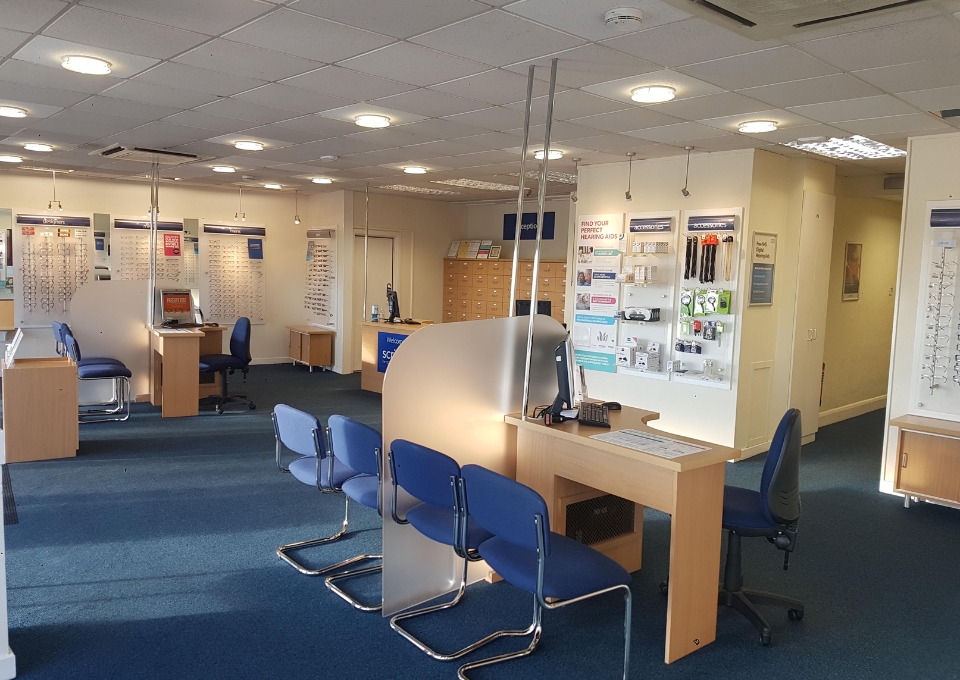 Bright optician's reception area with blue chairs, wooden desks, and wall display boards along the back wall.