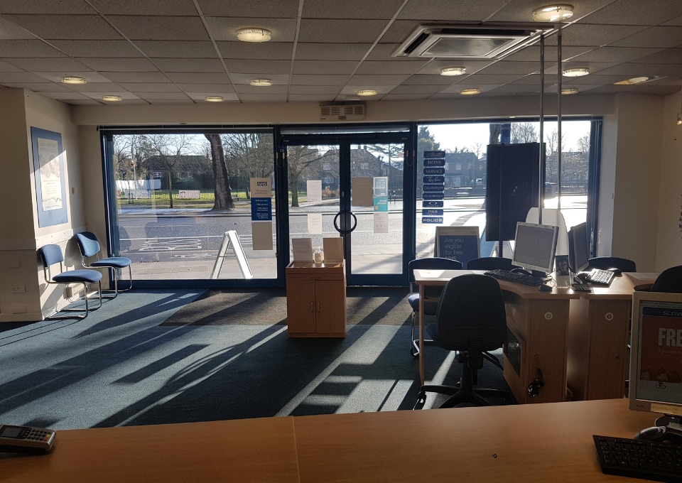 Reception area with glass front doors, blue chairs along the left wall, and a desk with computer on the right; outside street visible.