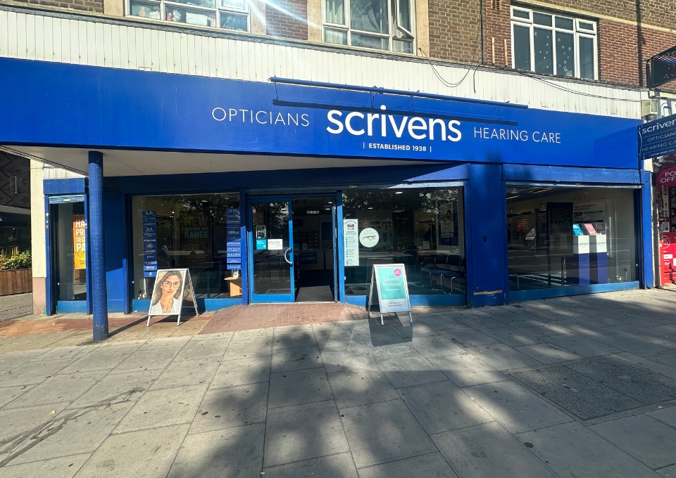 Blue storefront for Scrivens Opticians with Hearing Care signage on a sunny street. Glass doors and promotional boards outside the entrance appear on a bright day outside the shop.