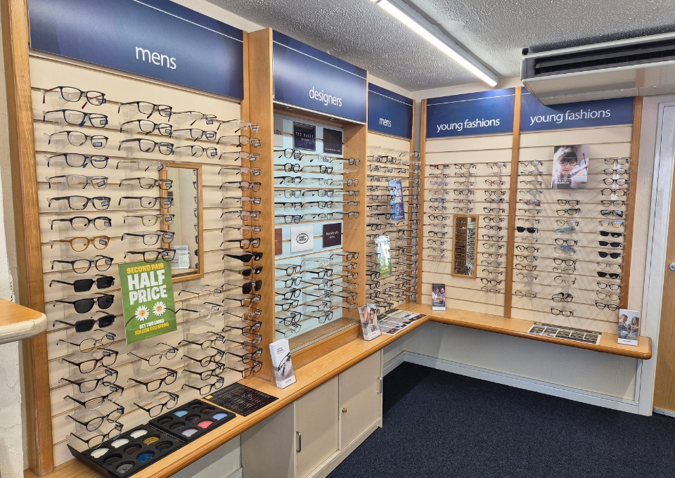 Interior of a eyewear store with wall-mounted racks displaying numerous glasses; blue signs read 'mens', 'designers', and 'young fashions' above sections.