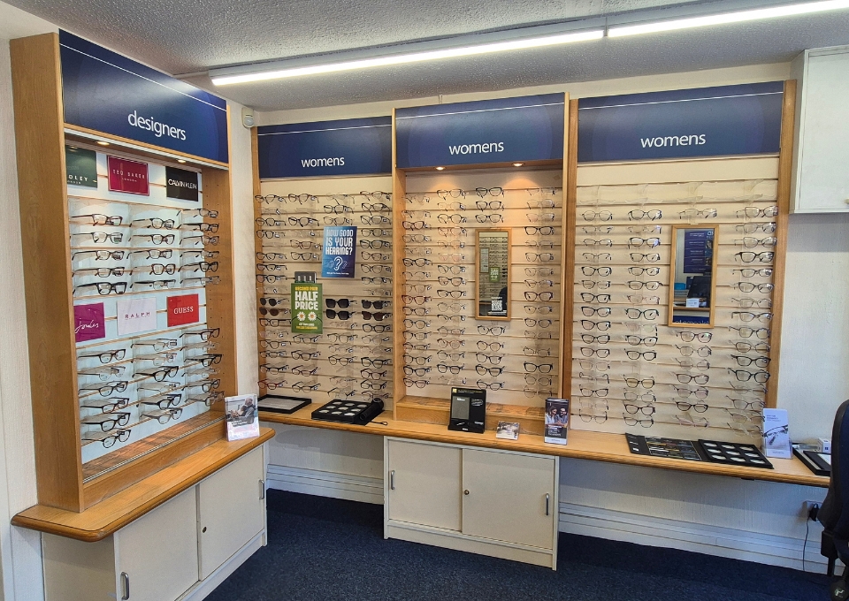 Wall display of women's designer eyeglasses in a retail optical shop, with frames arranged on panels and a wooden counter nearby
