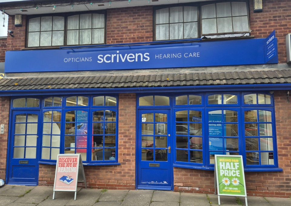 Blue Scrivens Opticians storefront with large blue sign, brick building, and blue-framed windows; two A-frame signs on the sidewalk show promotions.