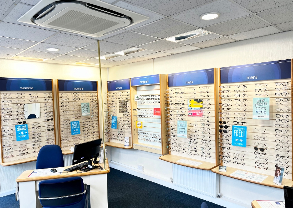 Eyewear shop interior with wall-mounted frames for women and men, a desk with a computer in the foreground.