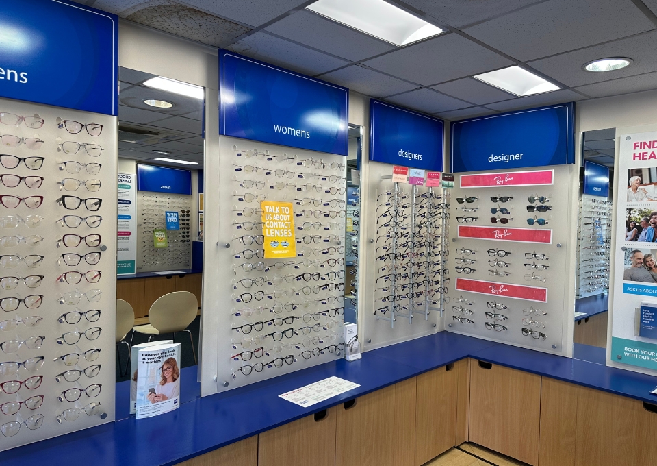 Eyeglasses on wall-mounted racks in an optical shop, with blue signs reading 'Womens' and 'Designer' above them and blue counters below.