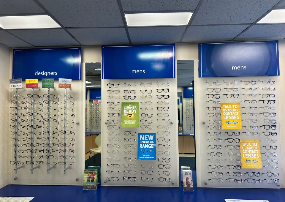 Eyewear display in a store with multiple racks of eyeglass frames under blue signs: 'designers' on the left and 'mens' in the center and right.