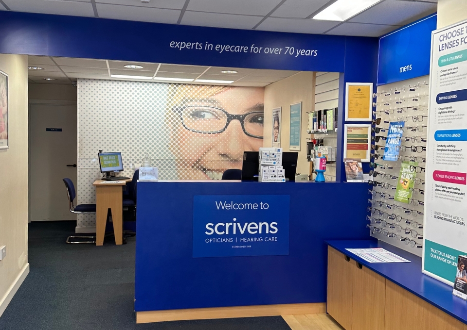 Reception area of Scrivens Opticians with a blue front desk, logo, and eyewear displays along the right wall. A large wall mural of a person wearing glasses sits behind the counter.