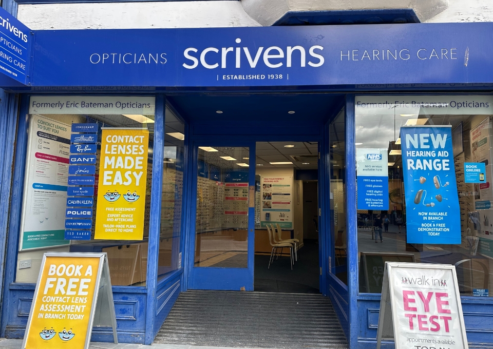 Blue Scrivens Opticians storefront with glass doors, blue signage, and posters advertising eye tests, contact lenses, and a free assessment offer