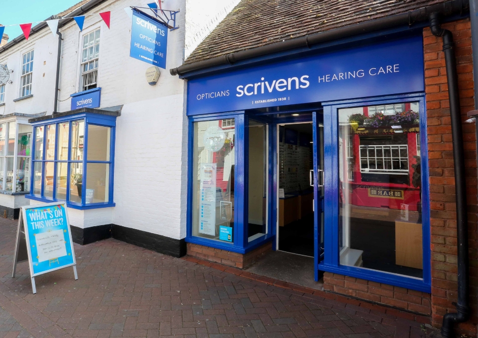 Blue-framed Scrivens hearing-care storefront with an open door, window displays, and a sidewalk signboard on a brick-paved street.