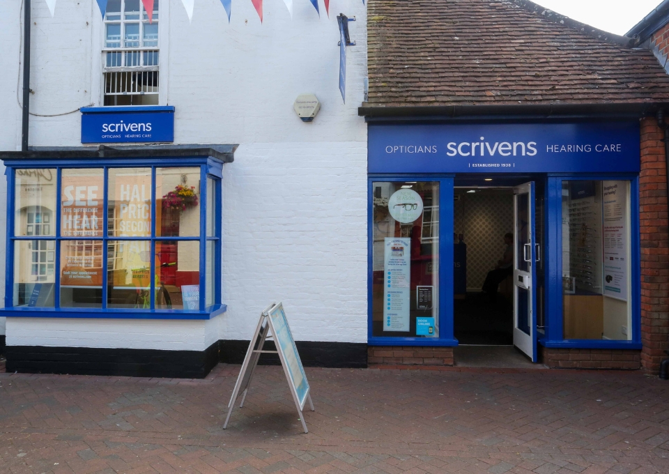 Blue-framed Scrivens opticians storefront with open door, window ads, and a sidewalk sandwich board on a brick-paved street.