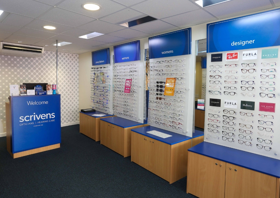 Store interior of Scrivens Opticians with wall displays of eyeglasses labeled women's, designer, and youth fashion sections on blue signs behind glass panels.