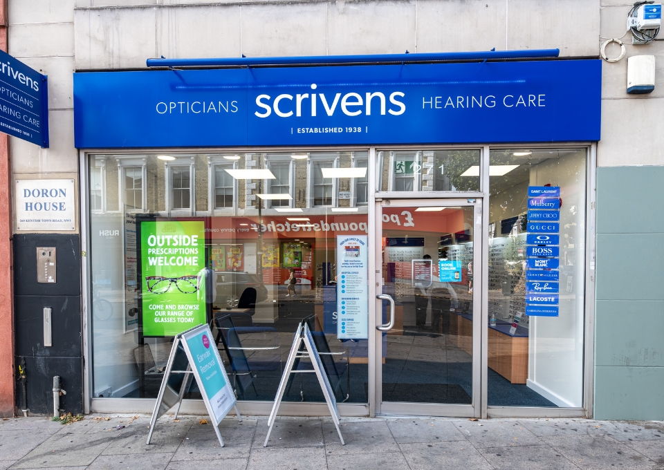 Storefront of an optician clinic with a blue sign reading 'Opticians scrivens HEARING CARE' across the top and glass doors below. Promotional posters are in the windows and on sidewalk signs outside the entrance.