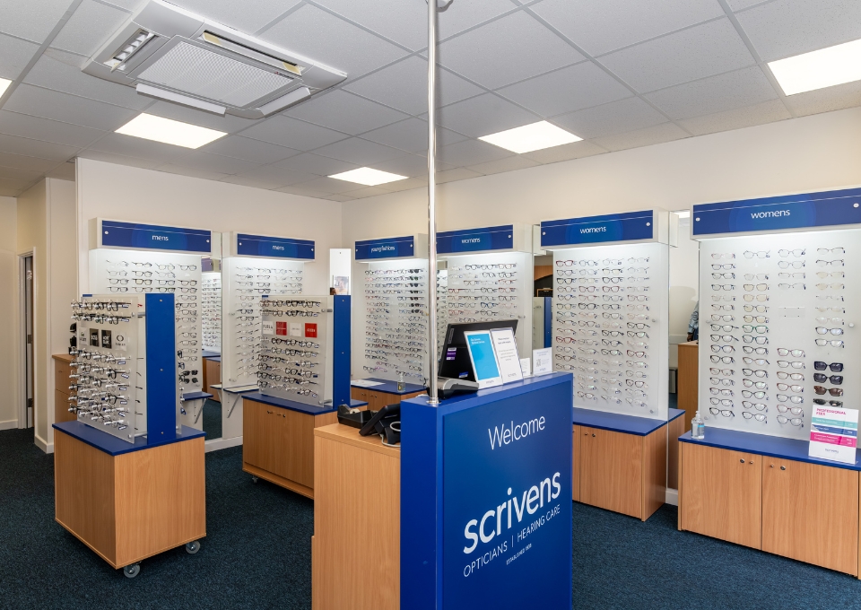 Interior of an eyewear store with wall displays of eyeglasses and a blue welcome counter labeled 'Scrivens Opticians.'