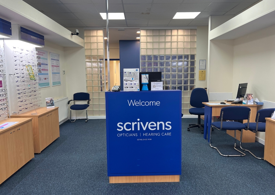 Blue reception desk in an eyewear store with 'Welcome scrivens' logo; glasses displays to the left and a seating area and computer to the right.