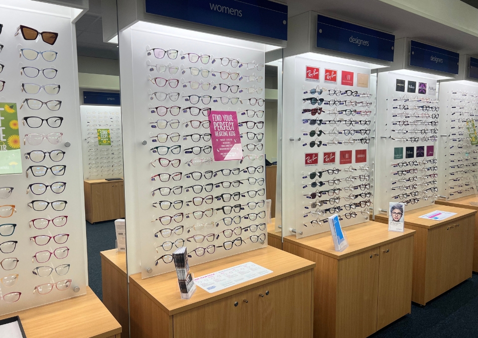 Wall of eyeglass frame displays in a store, with rows of frames on white panels and wooden counters underneath; blue signs read 'womens' and 'designers'.