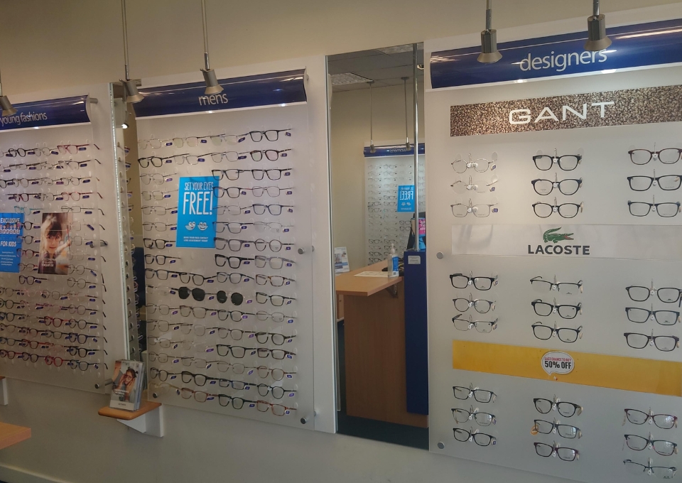 Eyeglasses displayed on wall panels in an eyewear store, with sections labeled young fashions, mens, and designers; GANT and Lacoste logos visible.