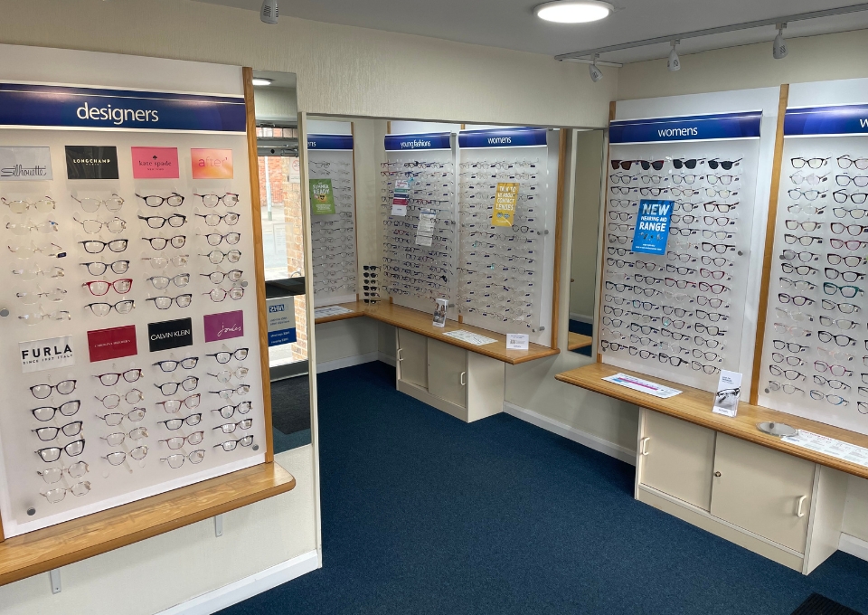 Eyewear store display with rows of eyeglasses on panels labeled 'designers' and 'womens' along wooden benches and white walls.