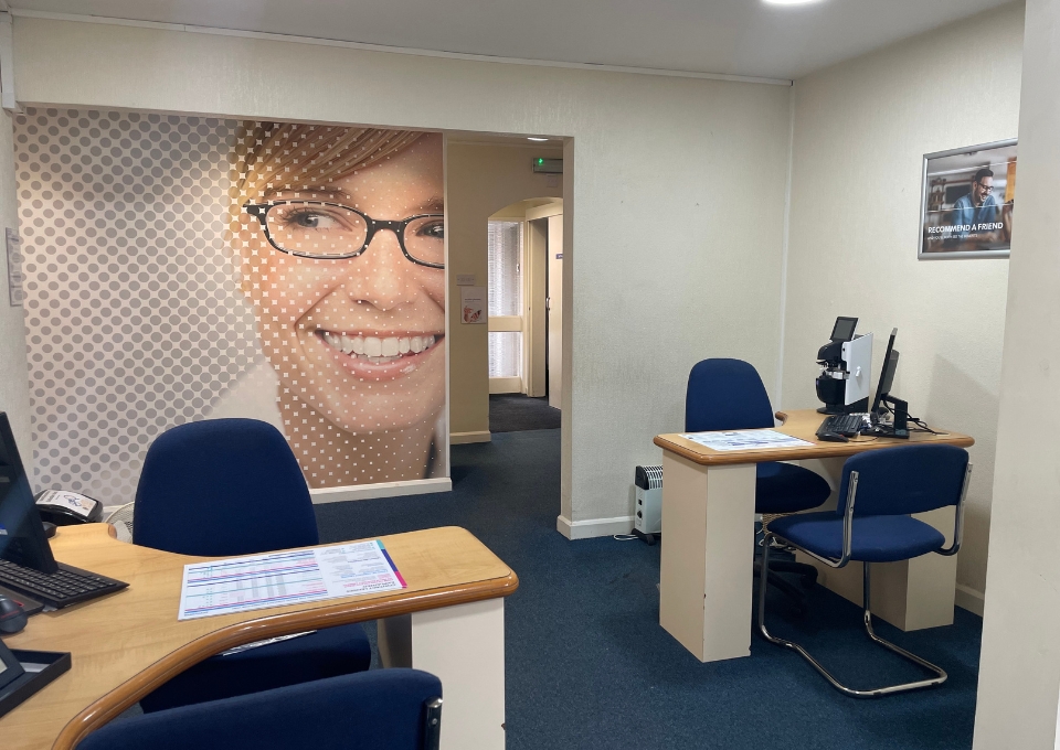 Office reception with two desks and computers; left wall has a large close-up of a smiling woman wearing glasses, with a dotted pattern backdrop.