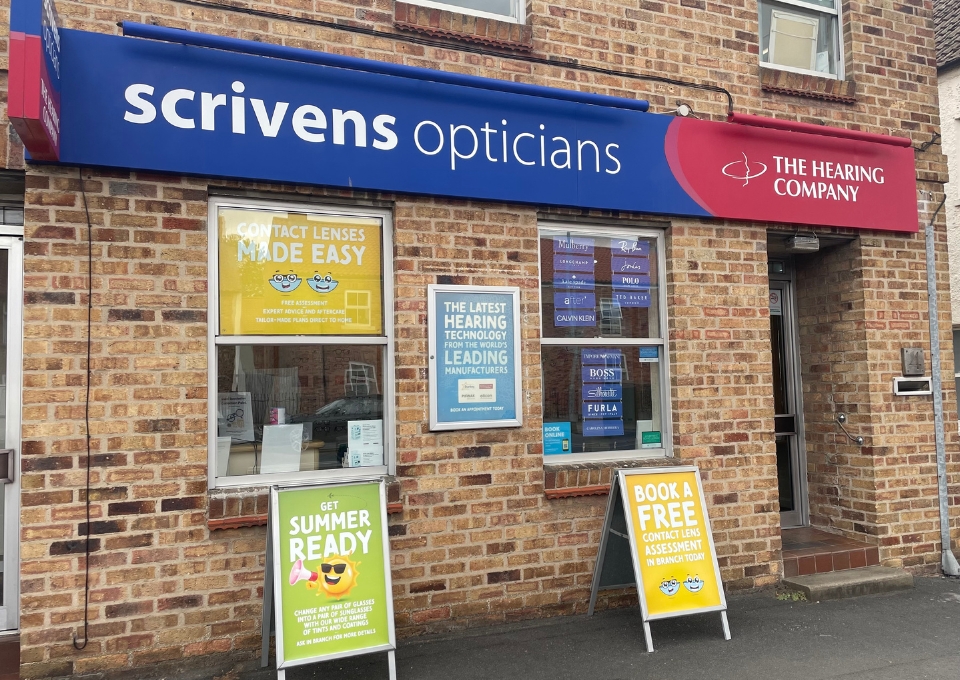 Blue Scrivens Opticians sign and red The Hearing Company panel on a brick storefront, with window posters advertising contact lenses and hearing technology; yellow 'Book a Free' sign outside.