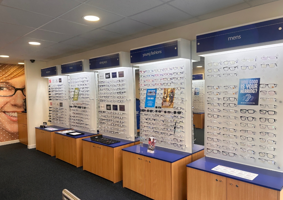 Wall of eyeglasses on display panels in an optical shop, labeled signs for men's, designers, and young fashions, with blue countertops beneath.