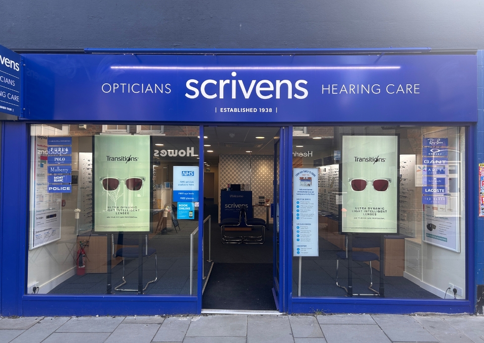 Blue storefront for Scrivens Opticians and Hearing Care with glass windows showing eyewear posters and an open entrance.