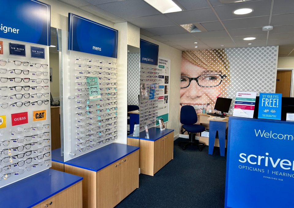 Interior of an optical store with display walls of prescription glasses labeled 'men's' and designer frames, blue accents, and a welcome desk reading 'scriv... Opticians' on the front panel.