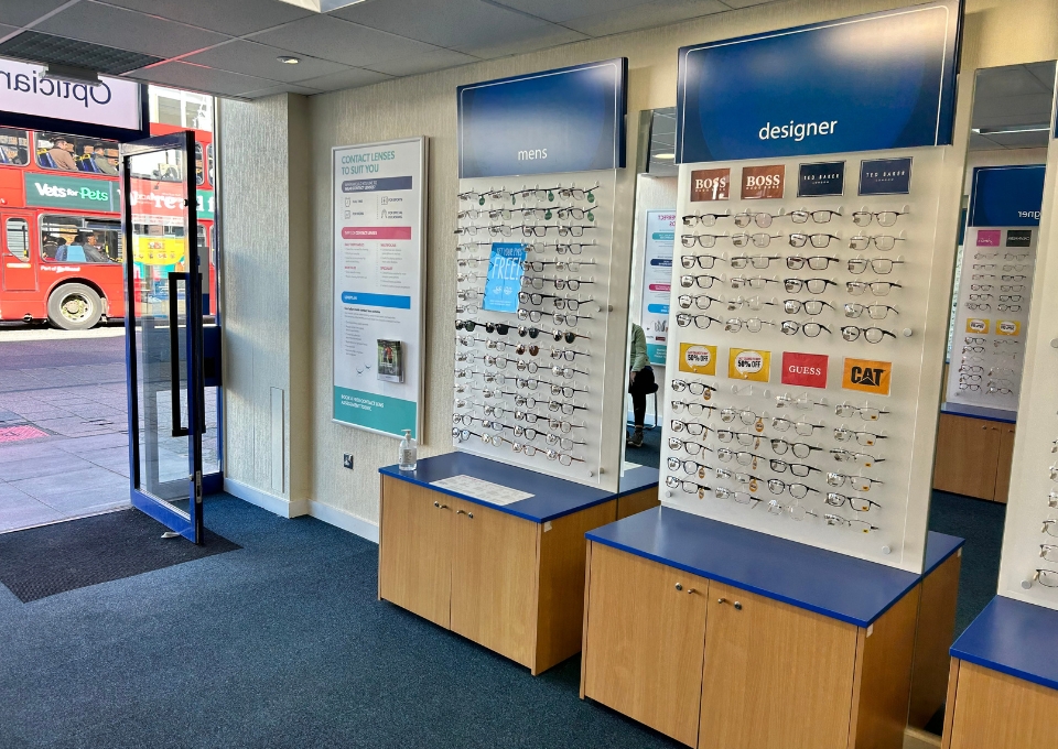 Interior of an eyewear shop with wall displays labeled 'mens' and 'designer' showcasing rows of eyeglasses on racks over wooden cabinets.