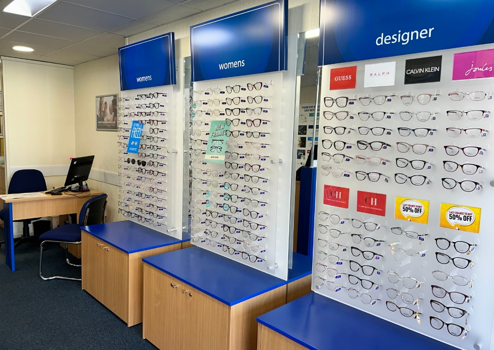 Store interior with wall-mounted eyewear displays labeled 'womens' and 'designer' showing rows of eyeglasses; blue signage, wooden storage units, and a desk with computer in the background