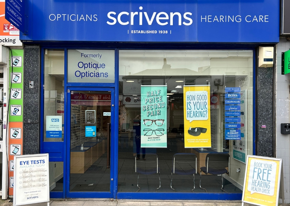 Blue storefront of Scrivens Opticians and Hearing Care with window posters promoting eye and hearing services.