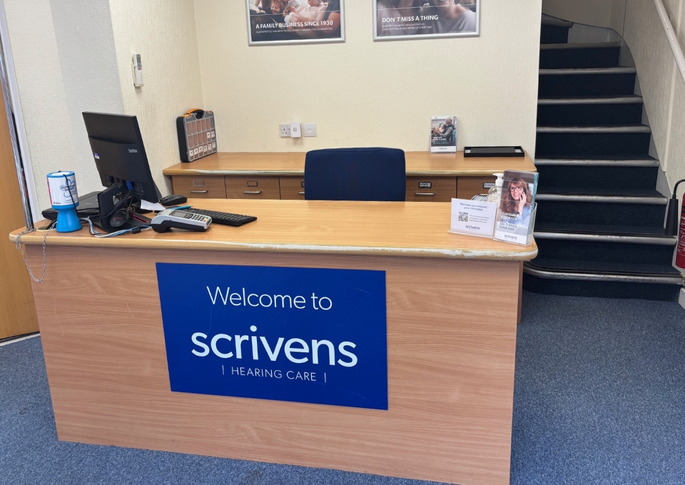 Reception desk at Scrivens Hearing Care with computer, phone, and brochures; staircase on the right.