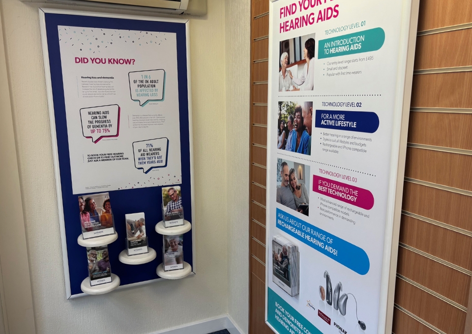 Two tall wall posters in a hallway: left board titled 'DID YOU KNOW?' with an infographic and four product displays on small round shelves; right board promoting hearing aids with 'FIND YOUR BEST HEARING AIDS' and technology levels.